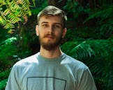 Portrait of a bearded man in a gray shirt, surrounded by lush green ferns in an outdoor setting.